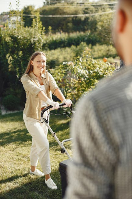 A woman with long brown hair, wearing a beige jacket, white trousers, and white sneakers, is engaged in garden maintenance, pushing a lawn mower across a well-kept grass lawn. The garden features lush