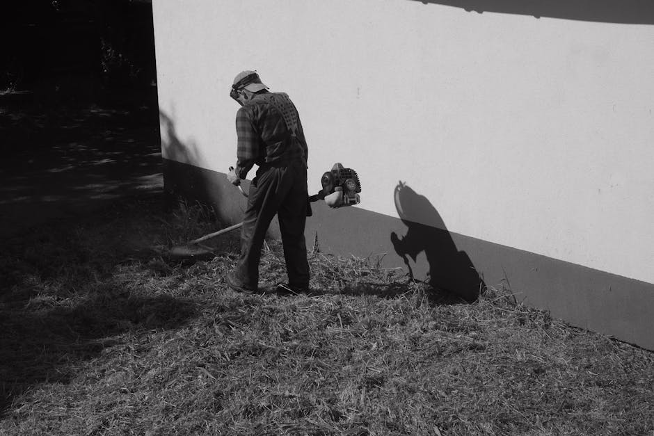 A woman with long brown hair, wearing a beige jacket, white trousers, and white sneakers, is engaged in garden maintenance, pushing a lawn mower across a well-kept grass lawn. The garden features lush